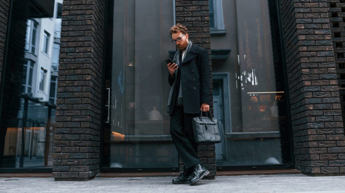 Holding smartphone. Stylish man with beard and in glasses is outdoors near building