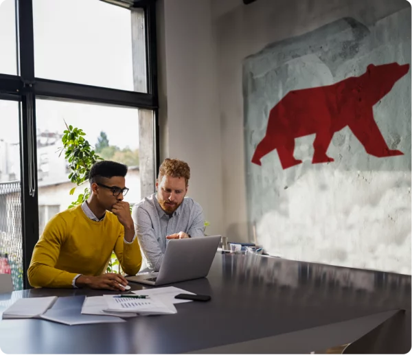 two men looking at a laptop in an office with Almas bear mural on the wall