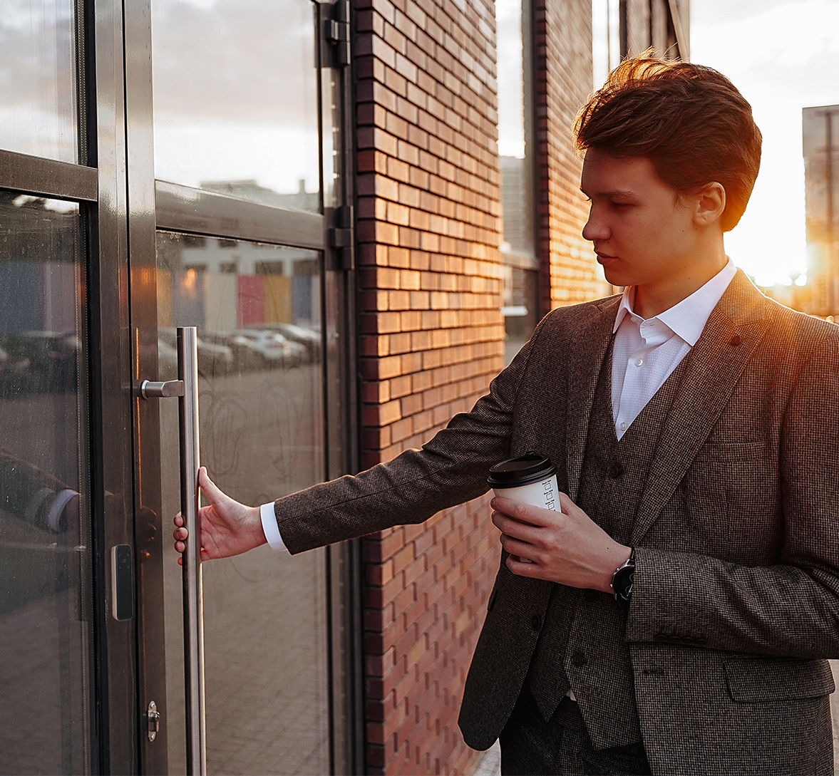 Young man in a suit holding a coffee opening a door with a fob reader on it.